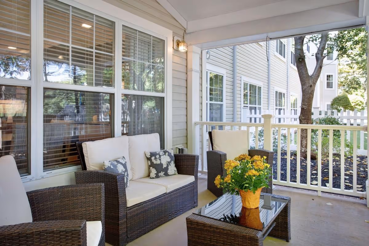 Covered outdoor patio with wicker seating, a glass-top coffee table holding a pot of yellow flowers, and a white railing overlooking landscaping.