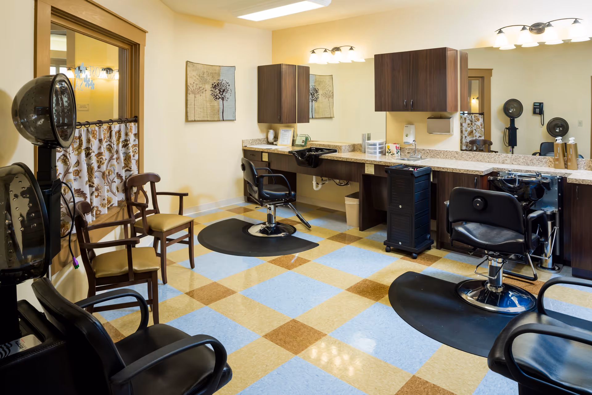 Interior view of a salon area in a senior living facility with two black salon chairs in front of sinks and mirrors, wooden cabinets above the sinks, two wooden chairs with cushions, two hair dryers, and a checkered floor with blue, beige, and brown tiles.