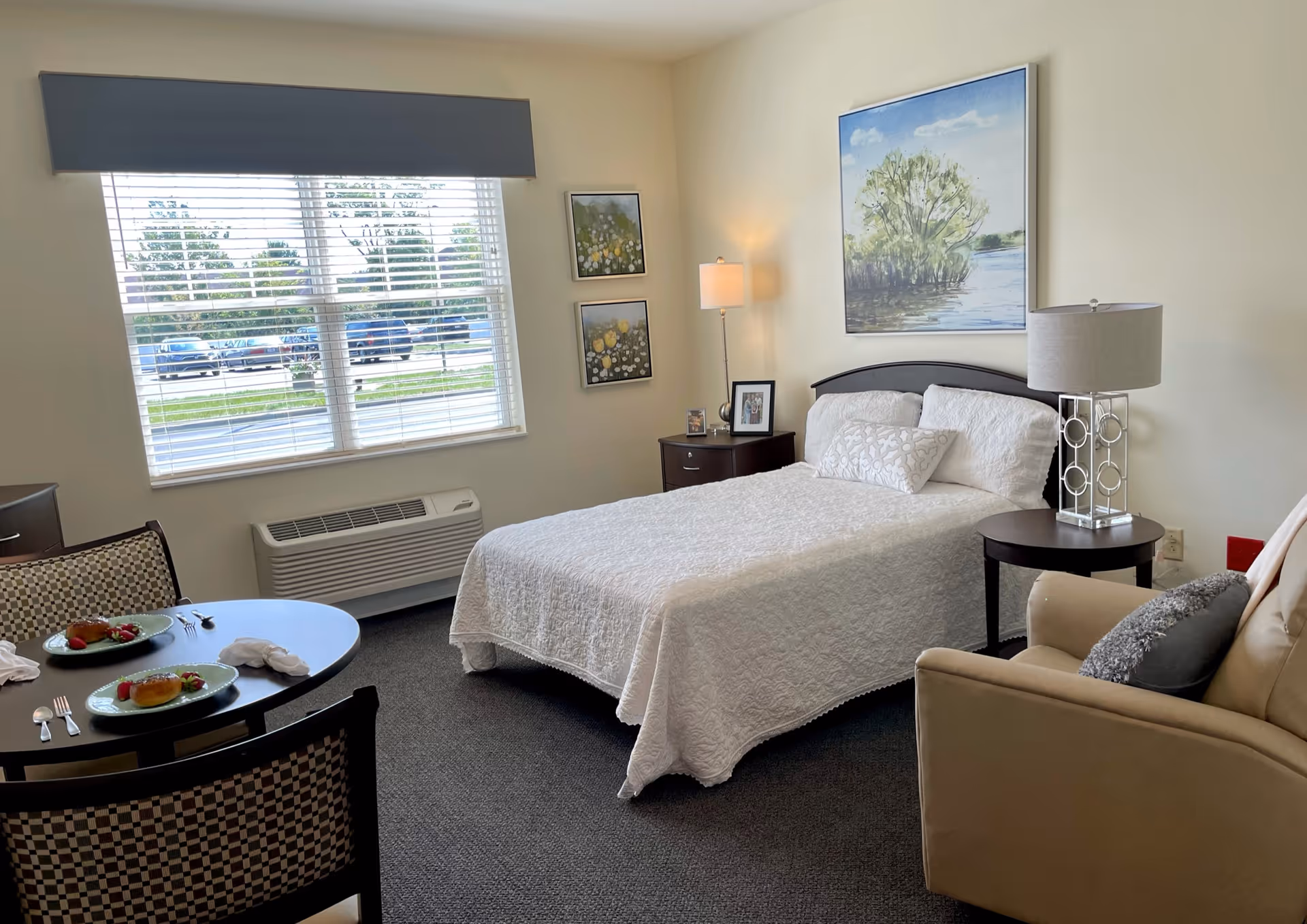 A cozy bedroom in a senior living facility with a neatly made bed covered in a white quilt. The room features a window with blinds, two framed floral pictures on the wall, a floor lamp, a side table with a lamp, and a comfortable armchair with a pillow. There is also a small round dining table set with two plates of food and utensils.
