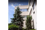 Exterior view of a multi-story white building facade with tall evergreen trees and a partly cloudy sky.