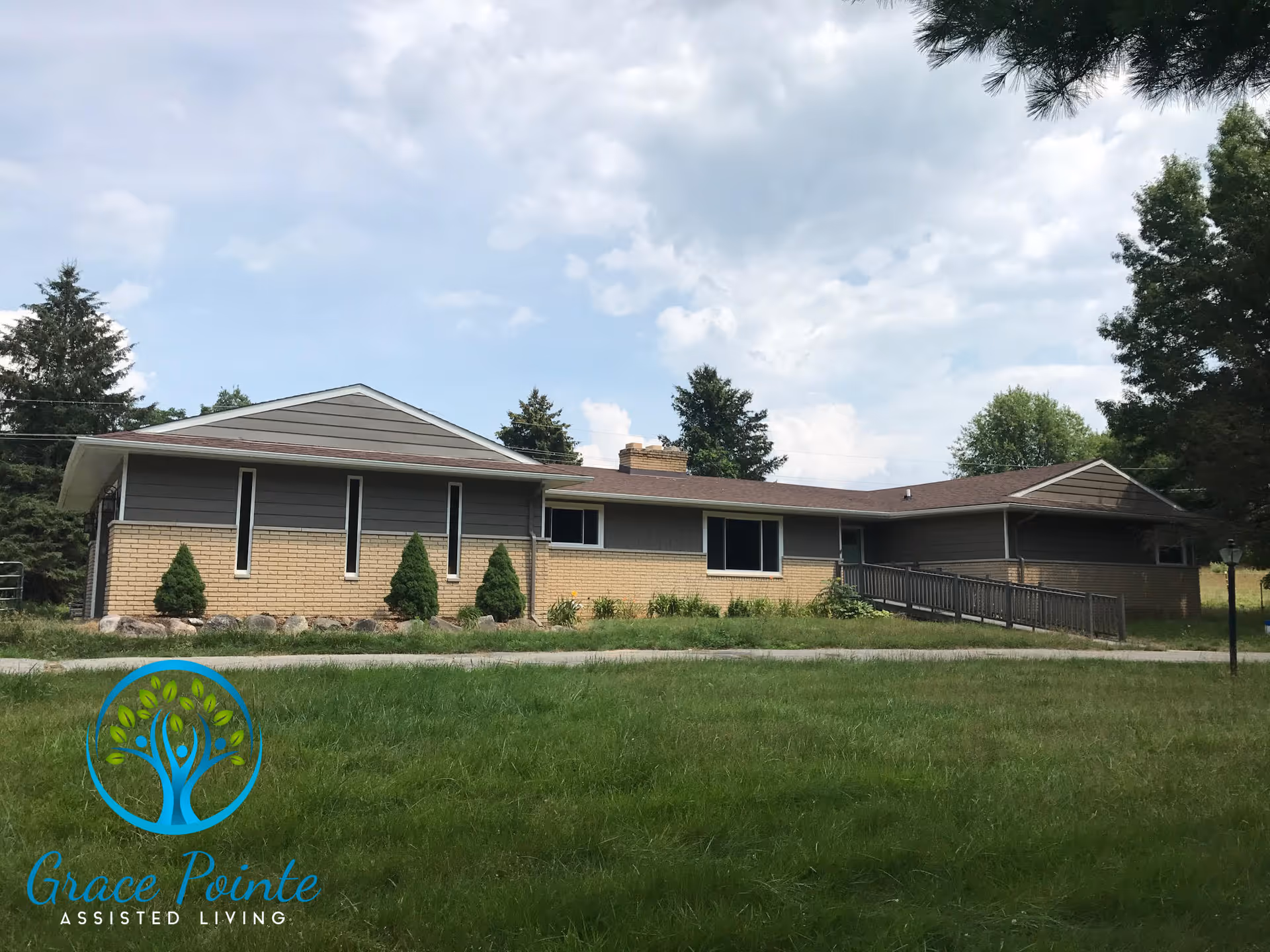 Single-story brick and wood assisted living building with a wheelchair ramp and front lawn under a cloudy sky.