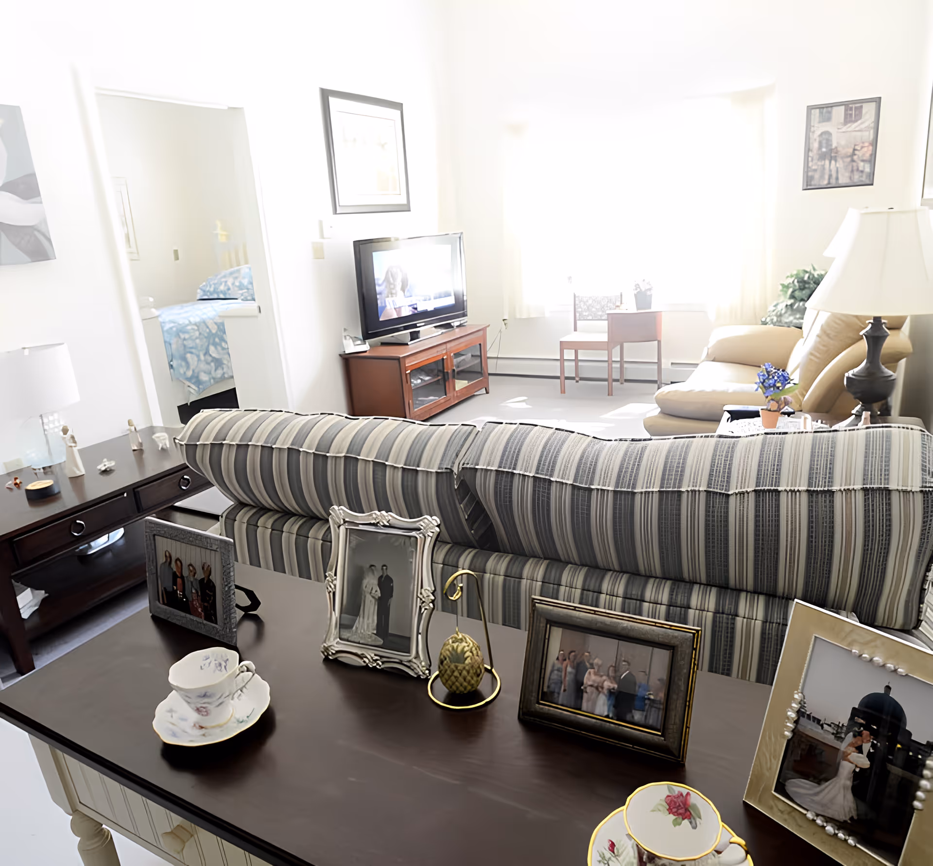 Bright living room with a striped sofa, TV and coffee table, a console table with framed photos and teacups in the foreground, and a bedroom visible through a doorway.
