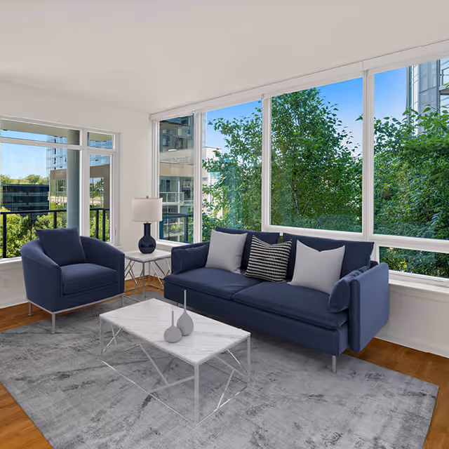 Bright living room with a navy sofa and armchair, a marble coffee table, and large windows overlooking trees.
