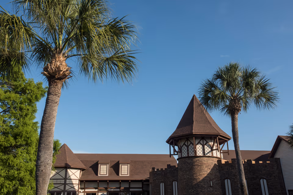 Exterior view of a building with castle-like architectural features, including a turret with a pointed roof, surrounded by palm trees under a clear blue sky.