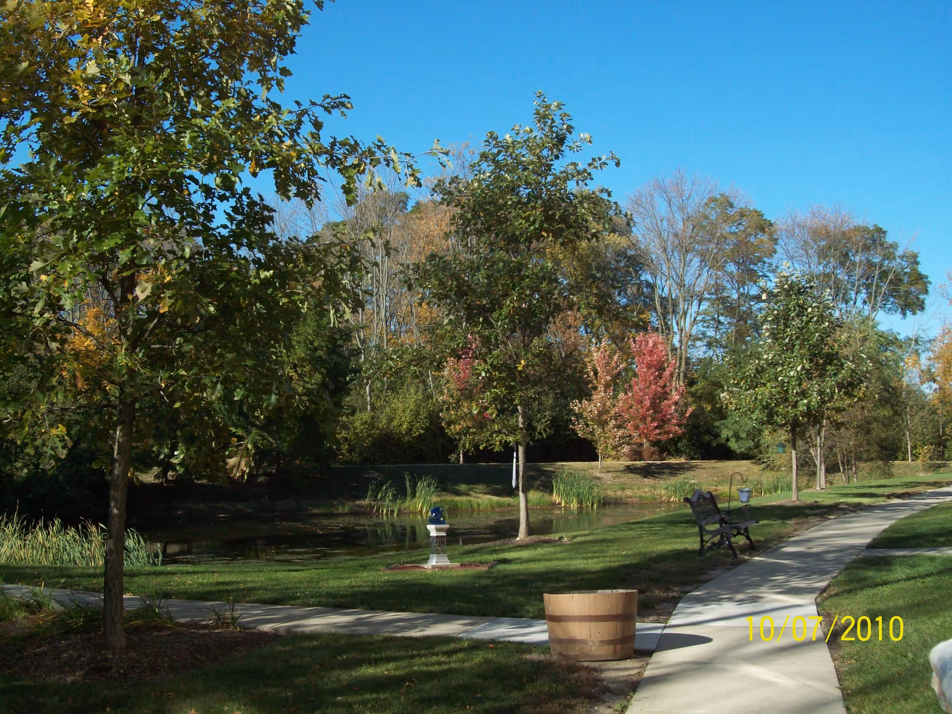 A peaceful outdoor scene at Lakewood Tower Apartments featuring a paved walkway curving through a grassy area with trees, a wooden barrel planter, a bench, and a small pond surrounded by greenery under a clear blue sky.