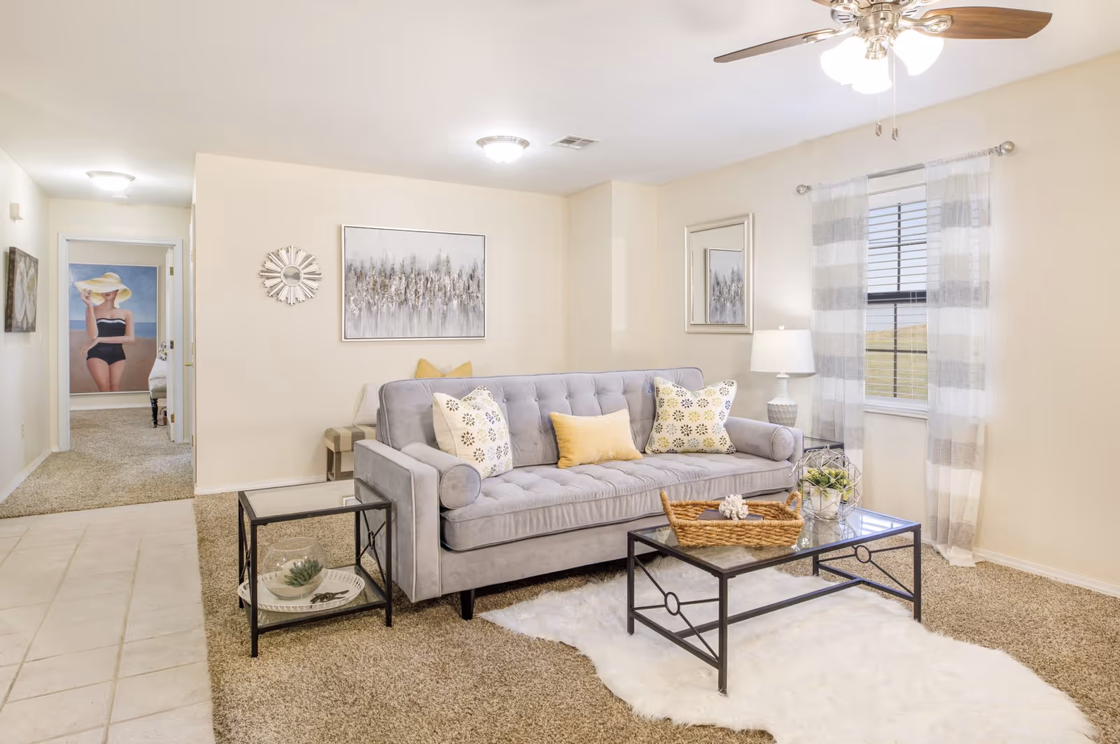A bright living room with a gray tufted sofa adorned with patterned and solid yellow pillows. In front of the sofa is a glass coffee table with a wicker tray and decorative items, placed on a white fluffy rug over beige carpet. To the left is a small glass side table with a decorative bowl and plant. The room has cream-colored walls, a ceiling fan with lights, and a window with sheer striped curtains. In the background, a hallway leads to another room with a large wall art of a woman in a black swimsuit and yellow hat.
