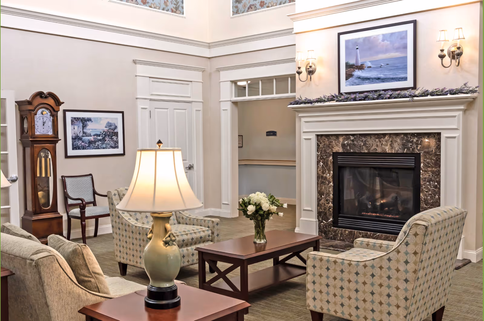 A cozy living room area in The Lighthouse at Lincoln featuring a fireplace with a marble surround and a framed picture of a lighthouse above it. The room has patterned armchairs, a sofa, a wooden coffee table with a vase of white flowers, and a table lamp. There is a grandfather clock and a framed landscape painting on the wall, with soft lighting from wall sconces.