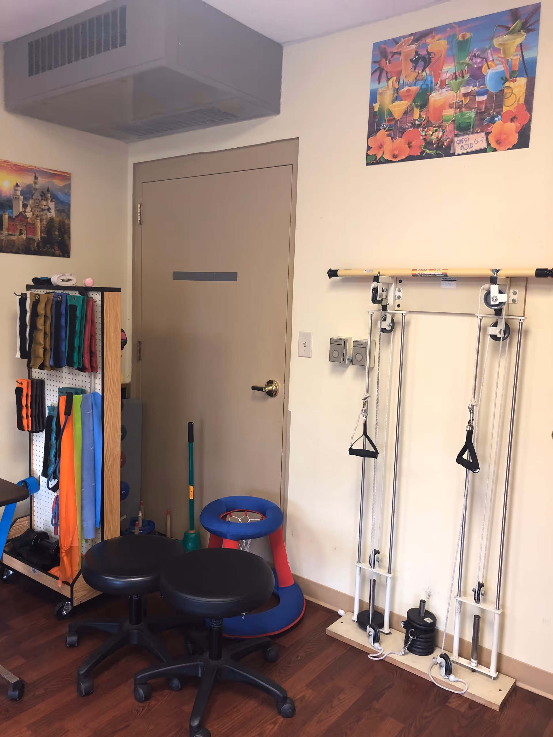Interior therapy room with exercise equipment, resistance bands, pulley machines and two black rolling stools in front of a closed beige door.