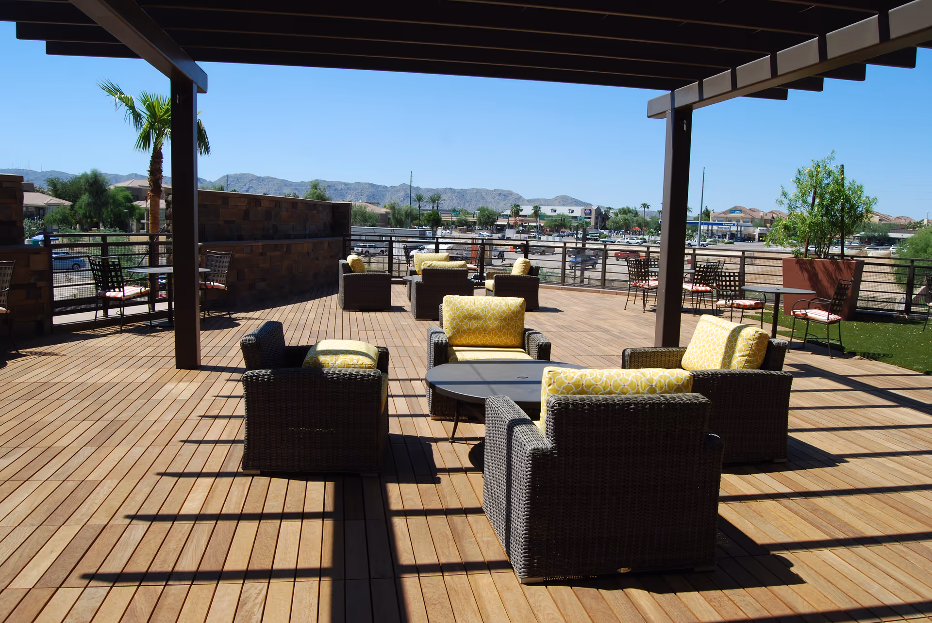 Outdoor patio area with wooden decking, featuring several wicker chairs with yellow cushions arranged around a low table. There are additional tables and chairs along the railing, potted plants, and a view of distant mountains under a clear blue sky.