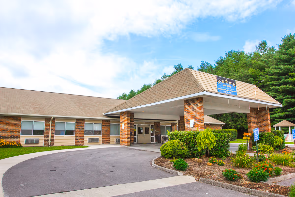 Exterior view of a single-story brick building with a covered entrance supported by brick columns. There is a circular driveway in front and landscaped bushes and flowers around the entrance. The building is surrounded by trees and under a partly cloudy sky.