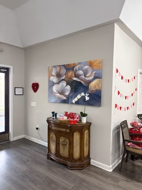 Interior corner of a room with a wooden cabinet against a beige wall. Above the cabinet is a large floral painting featuring white and orange flowers. On the cabinet are decorative items including a small potted orchid and other small objects. To the right, part of a dining table with chairs is visible, decorated with red and white garlands hanging on the adjacent wall. The floor is dark wood, and a door with a window is visible on the left side.