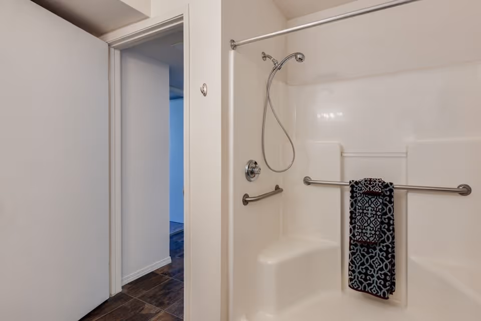 Interior view of a bathroom shower area with built-in seating and grab bars. A patterned towel is hanging on one of the grab bars. The shower has a handheld showerhead attached to a flexible hose. The bathroom door is open, showing a hallway with dark wood flooring.