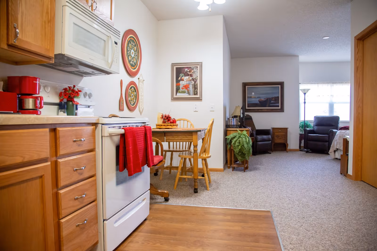 Interior view of a senior living facility showing a kitchen area with wooden cabinets, a white stove with a red towel hanging on it, a microwave, and a red coffee maker. Adjacent to the kitchen is a small dining area with a wooden table and chairs. Beyond the dining area is a living space with two recliner chairs, a side table, a floor lamp, and framed artwork on the walls. The flooring transitions from wood in the kitchen to carpet in the living and dining areas.