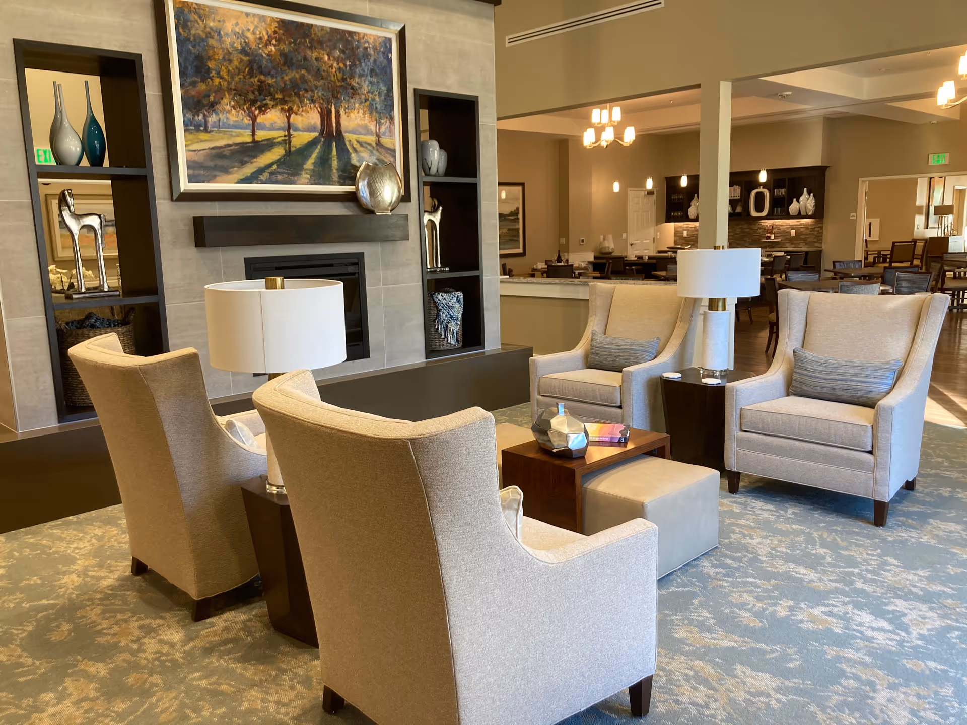 A cozy living room area in an assisted living facility featuring four beige armchairs arranged around a wooden coffee table with decorative items. The room has a fireplace with built-in shelves on either side displaying vases and sculptures. A large painting of trees hangs above the fireplace. In the background, there is a dining area with tables and chairs, and warm lighting fixtures on the ceiling.