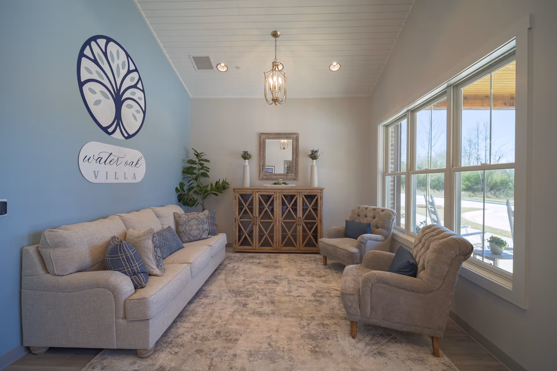 A cozy sitting area in Water Oak Villa featuring a beige sofa with multiple patterned cushions, two matching armchairs with blue pillows, a wooden cabinet with glass doors topped with two vases and a mirror, a large window letting in natural light, and a blue accent wall with the Water Oak Villa logo.