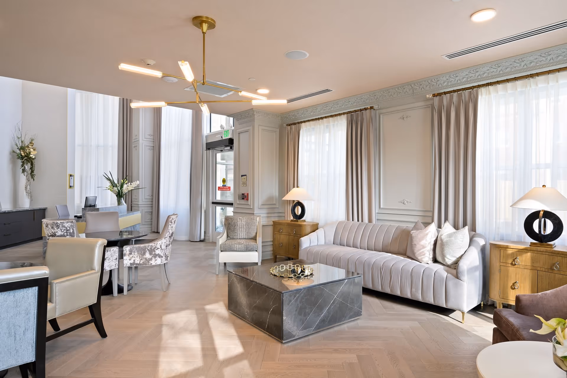 A bright and elegant living room area with a modern gray sofa adorned with pillows, two wooden side tables with black circular lamps, a square marble coffee table, and several upholstered chairs around a small round table. Large windows with sheer and beige curtains allow natural light to fill the room. The space features light wood flooring, decorative wall molding, and a contemporary gold chandelier hanging from the ceiling.