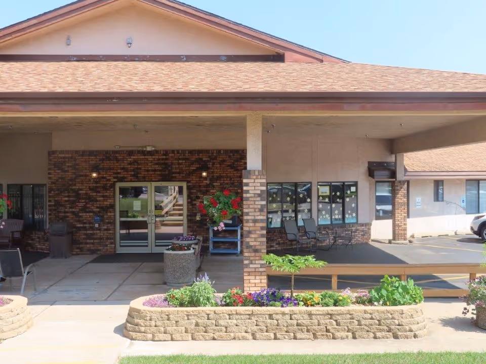Entrance of a senior living facility with a covered porch area, brick and beige exterior walls, glass double doors, potted plants, outdoor chairs, and a flower bed in front.