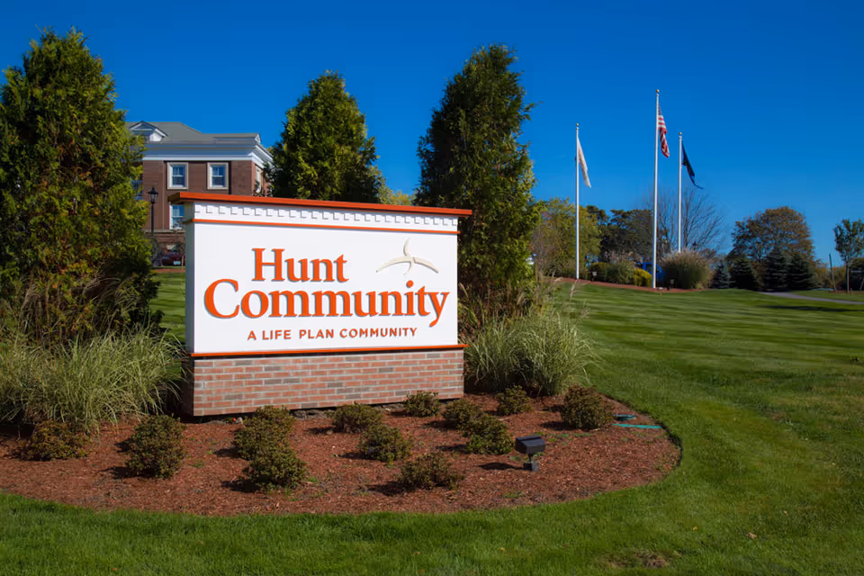 Outdoor view of the Hunt Community sign on a landscaped lawn with bushes and trees, with a building and three flagpoles flying flags in the background under a clear blue sky.