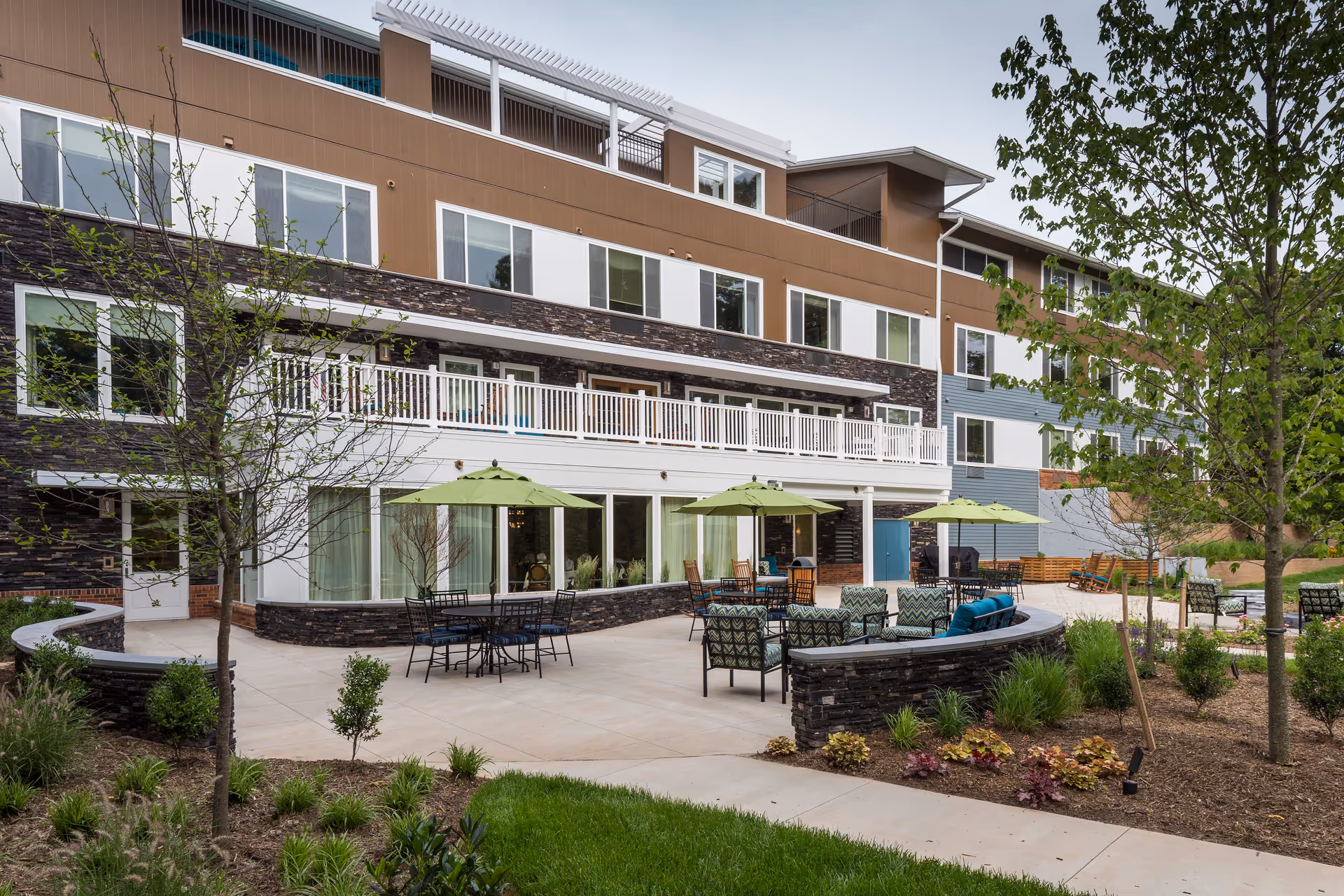 Outdoor patio area of a senior living facility with multiple seating arrangements including tables with green umbrellas and cushioned chairs. The patio is surrounded by landscaped greenery and trees, and the building has multiple floors with large windows and a balcony.