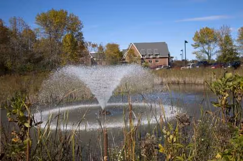 A pond with a fountain and a brick building in the background.