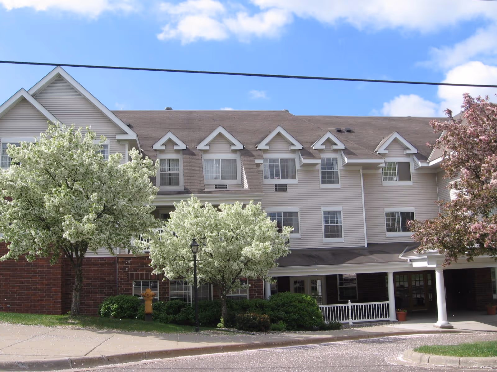 Exterior view of a multi-story senior living facility building with beige siding and brown roof, surrounded by blooming white and pink flowering trees under a blue sky with some clouds.