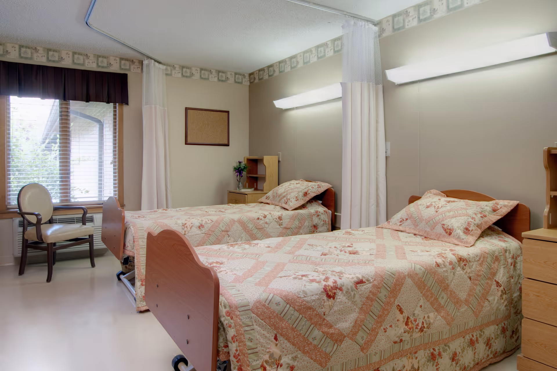 A room with two single beds covered in floral patterned quilts. Each bed has a pillow and a wooden headboard. There is a small wooden nightstand between the beds with a vase of flowers on it. A chair is placed near a window with blinds and a dark valance. The walls are light-colored with a decorative border near the ceiling, and there are two long fluorescent lights mounted above the beds. Privacy curtains hang from the ceiling between the beds.