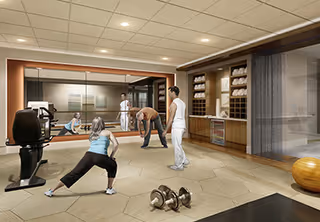 A fitness room in a senior living facility with exercise equipment, including dumbbells and a stationary bike. Four people are engaged in stretching and exercise activities, with one instructor guiding them. The room has a large mirror on one wall, shelves with towels, and a warm, inviting atmosphere.