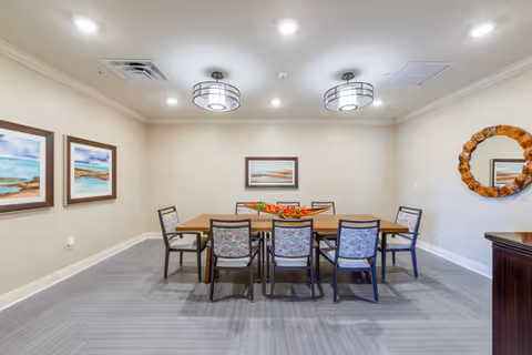 A dining room with a rectangular wooden table surrounded by eight chairs with patterned cushions. The walls are light beige and decorated with three framed landscape paintings and a round wooden-framed mirror. The ceiling has two modern circular light fixtures and recessed lighting. The floor is covered with gray tiles.