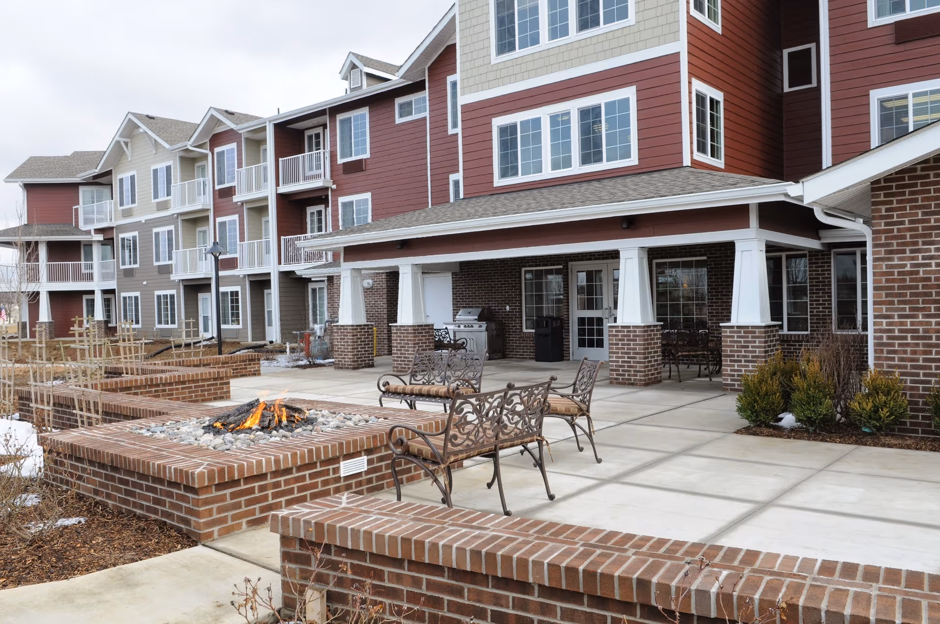 Outdoor patio area of a senior living facility with brick fire pits, metal benches with cushions, and a covered seating area. The building has multiple floors with balconies and a mix of red, beige, and gray siding.