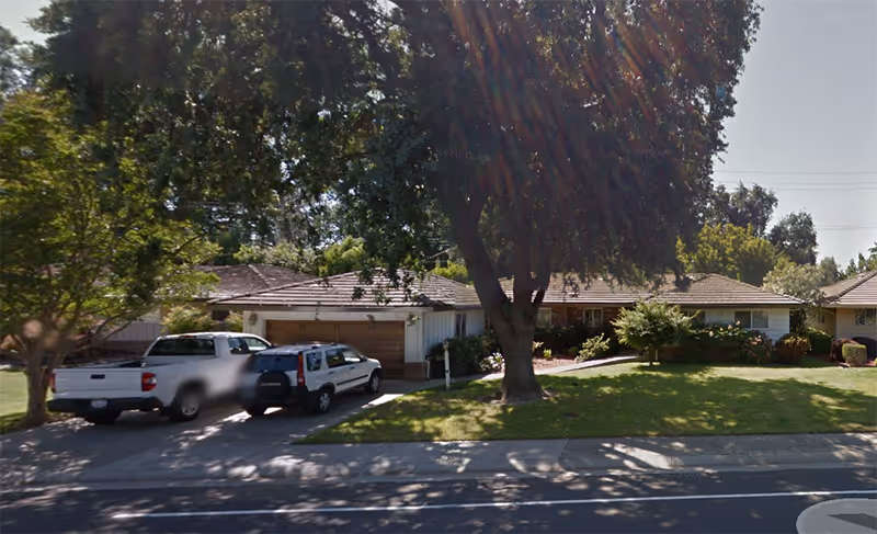 Front exterior of a single-story home with a driveway, trees, and parked vehicles.