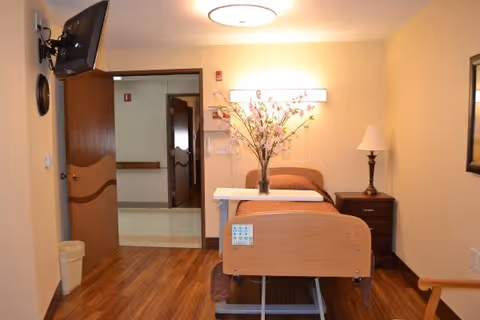 A long term care unit room with a single hospital bed centered against the back wall. The bed has a wooden headboard and footboard, with a vase of flowers placed on a tray on the bed. To the right of the bed is a wooden nightstand with a lamp. The room has wood flooring and beige walls. A door is open to a hallway, and a wall-mounted TV is visible on the left side.