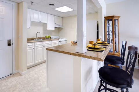 A bright kitchen area in a retirement community with white cabinets, a countertop with a sink, and a stove. There is a breakfast bar with three black cushioned chairs set with plates and cups. A glass display cabinet is visible in the background.