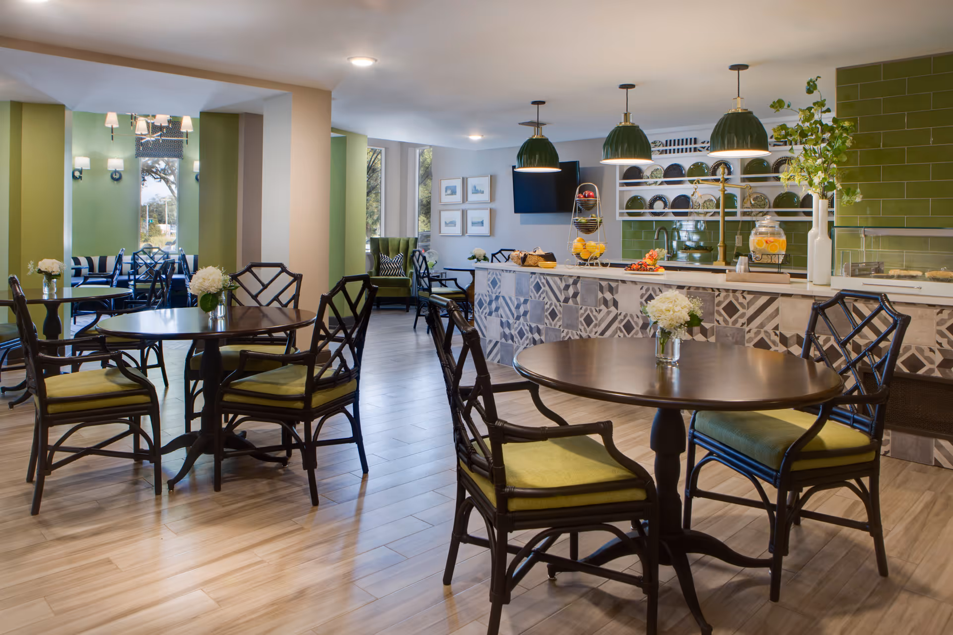 A bright and modern dining area with round wooden tables and dark wooden chairs with green cushions. The room features a tiled counter with decorative patterns, green tiled backsplash, and three green pendant lights hanging above. There are plates displayed on shelves behind the counter, a beverage dispenser with lemon water, and a few floral arrangements on the tables. Large windows allow natural light to enter, and there is a green armchair in the background near framed pictures on the wall.