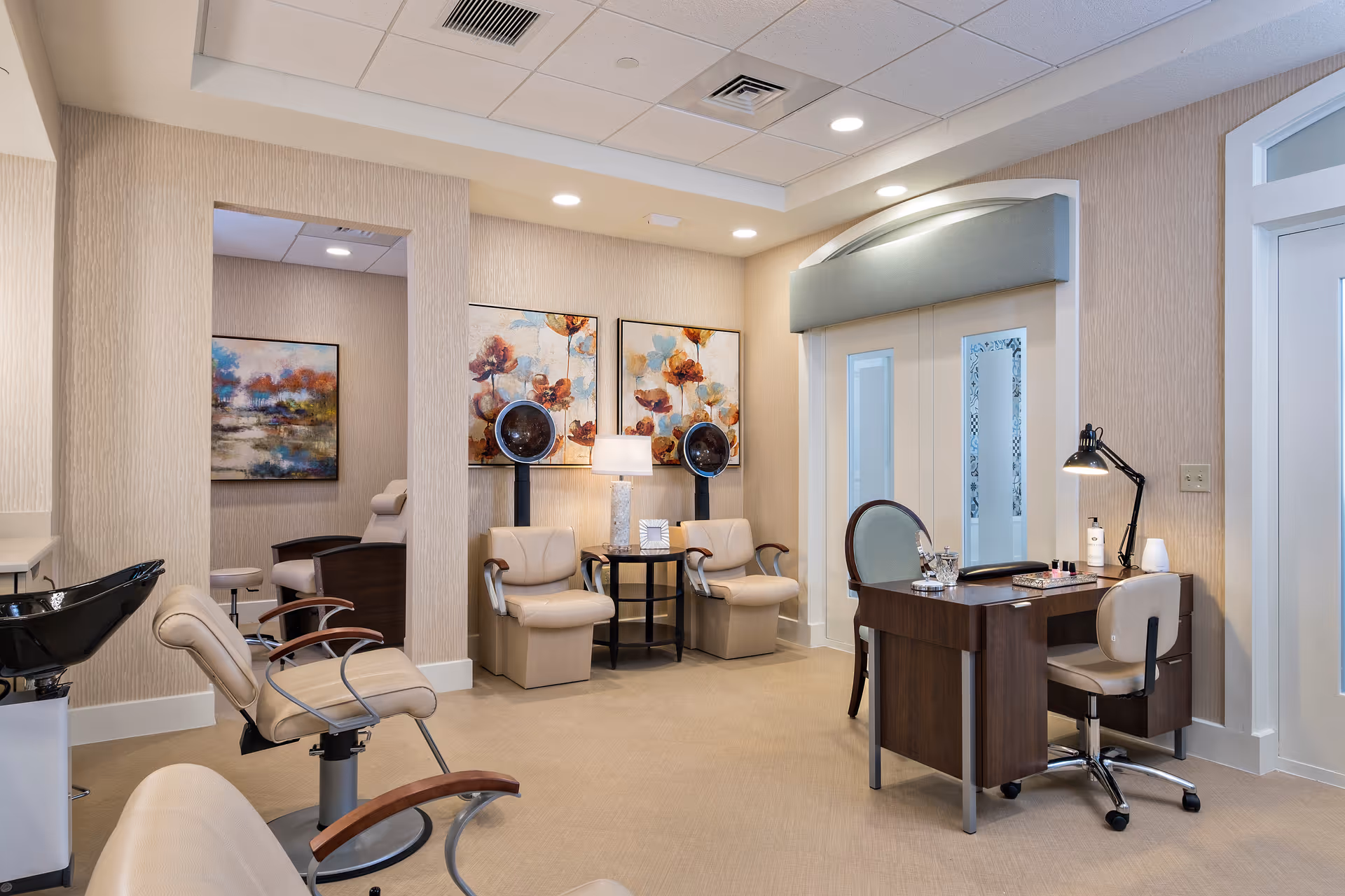 Interior view of a salon area in a senior living facility with beige walls and carpet. The room features salon chairs, hair dryers, a washing station, a desk with a lamp, and decorative paintings on the walls.