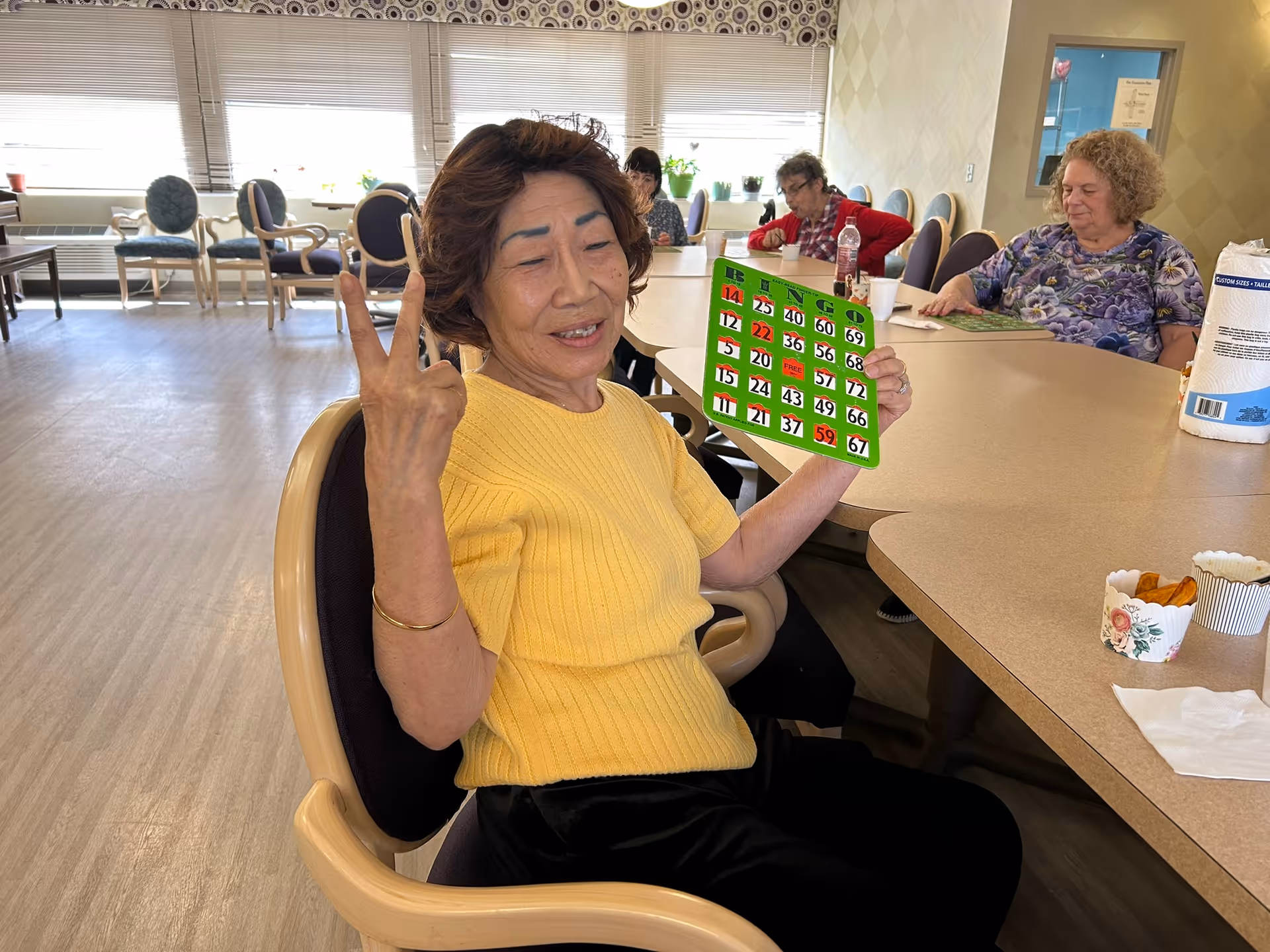 An elderly woman in a yellow sweater sitting at a table in a communal room, holding up a bingo card and making a peace sign with her fingers. Other elderly individuals are seated around the table, engaged in activities. The room has large windows with blinds and several chairs arranged along the walls.