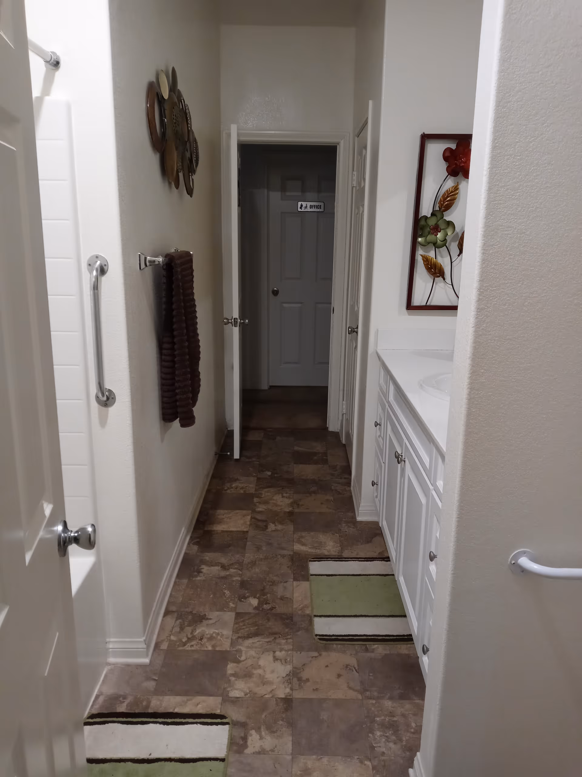A narrow bathroom hallway with tiled floor and white walls. On the left side, there is a bathtub with a grab bar and a towel hanging on a rack. On the right side, there is a white vanity with a sink and a decorative floral wall art above it. Two striped rugs are placed on the floor. At the end of the hallway, a door is open, revealing another door labeled 'Office'.