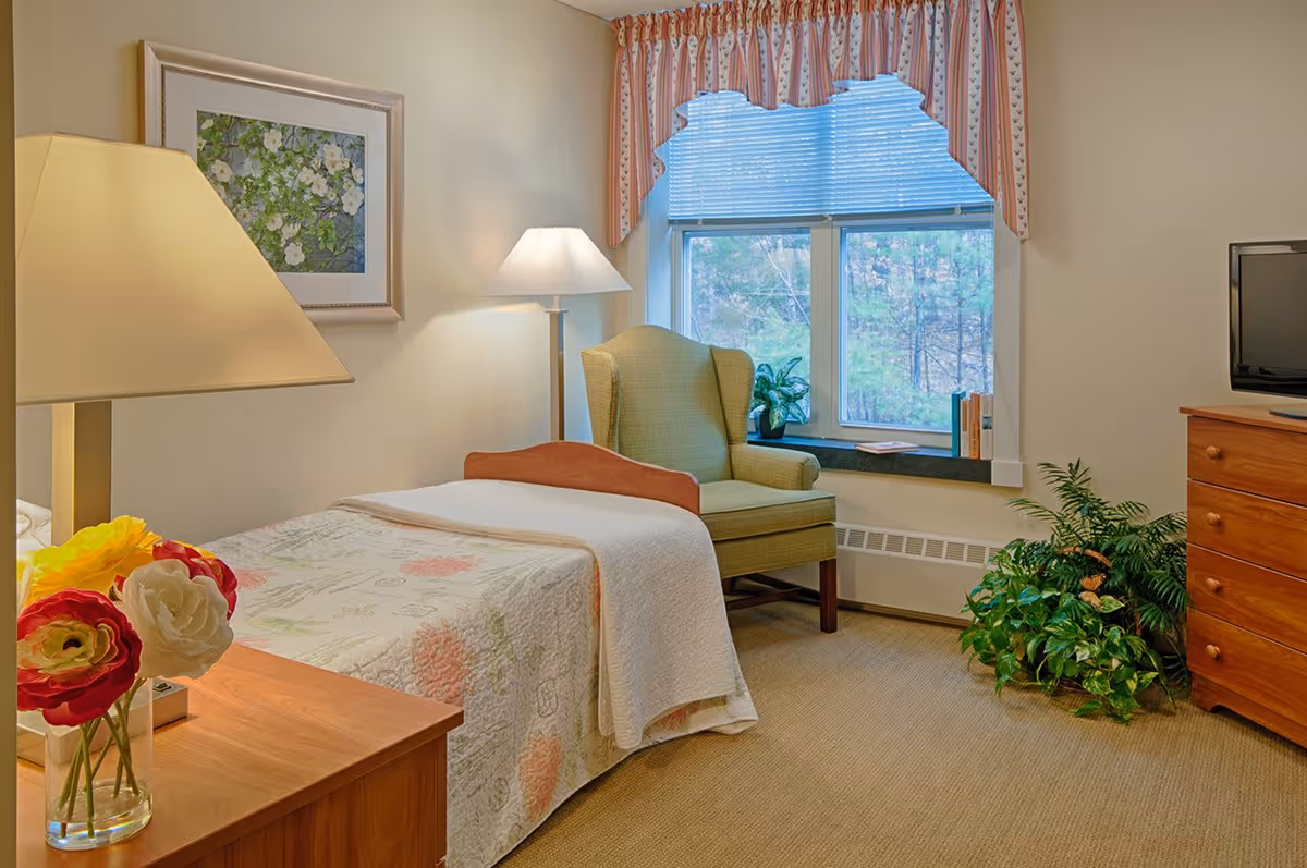 A cozy bedroom in a senior living facility with a single bed covered in a floral quilt, a wooden nightstand with a lamp and a vase of flowers, a green upholstered armchair near a window with blinds and a valance, a wooden dresser with a TV on top, and several green plants on the floor.