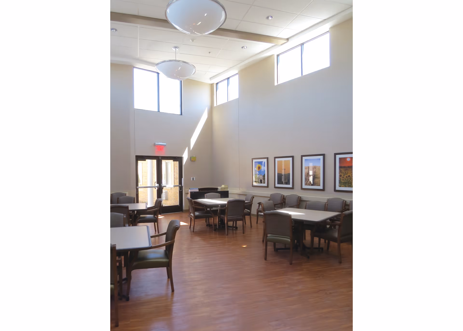 A bright, spacious room with high ceilings and large windows letting in natural light. The room contains several tables with chairs arranged around them, and framed pictures hanging on the wall. The floor is wooden, and there is a double glass door at the far end of the room.