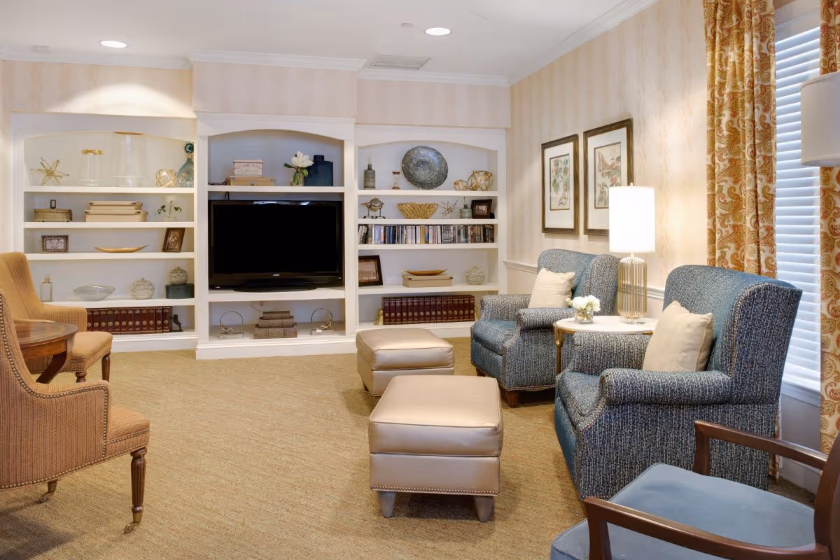 A cozy living room with two blue upholstered armchairs, two beige ottomans, a wooden chair, and a round wooden table. The room features built-in white shelves filled with decorative items, books, and a flat-screen TV. There are two framed pictures on the wall, a table lamp, and patterned curtains on the window.