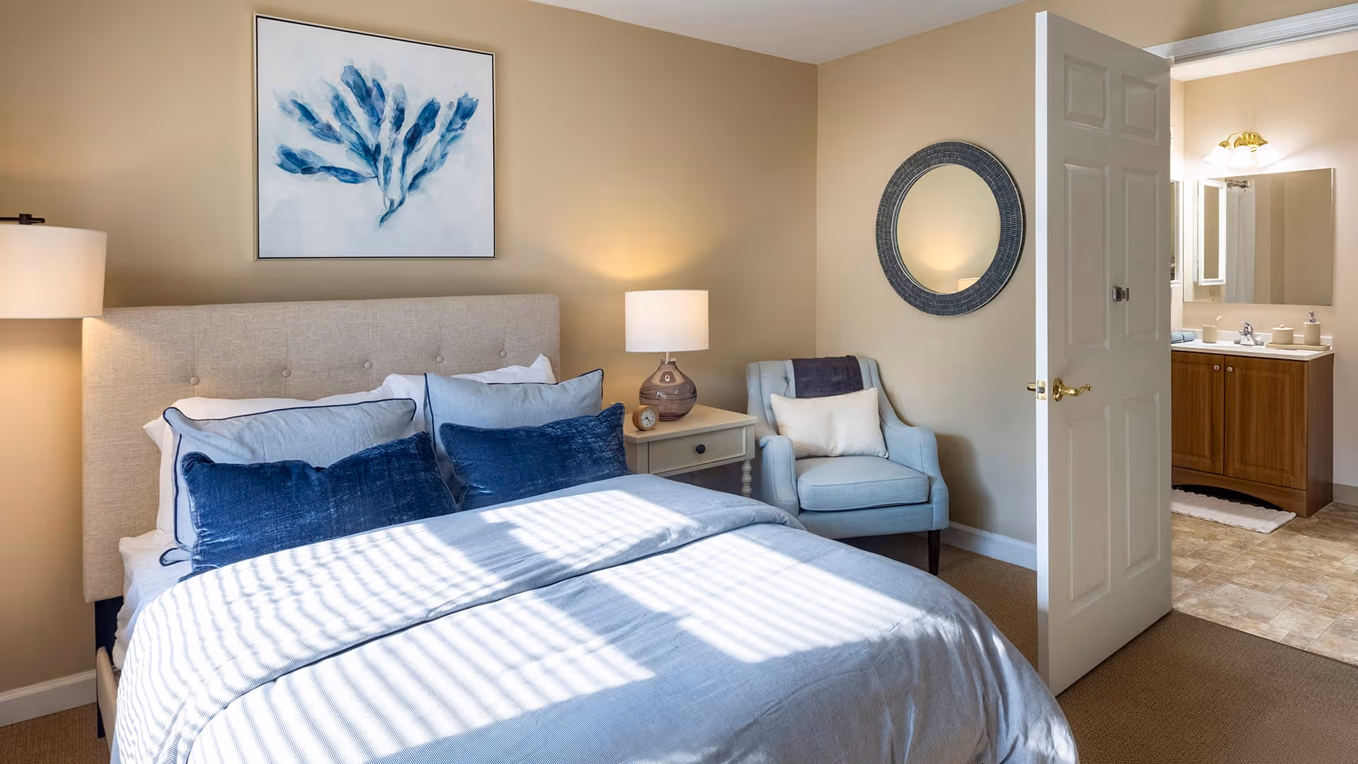 A cozy bedroom with a beige upholstered headboard, a bed made with light blue and navy blue pillows and bedding. There is a nightstand with a lamp and clock, a light blue armchair with a pillow, a round mirror on the wall, and an open door leading to a bathroom with a sink and mirror.