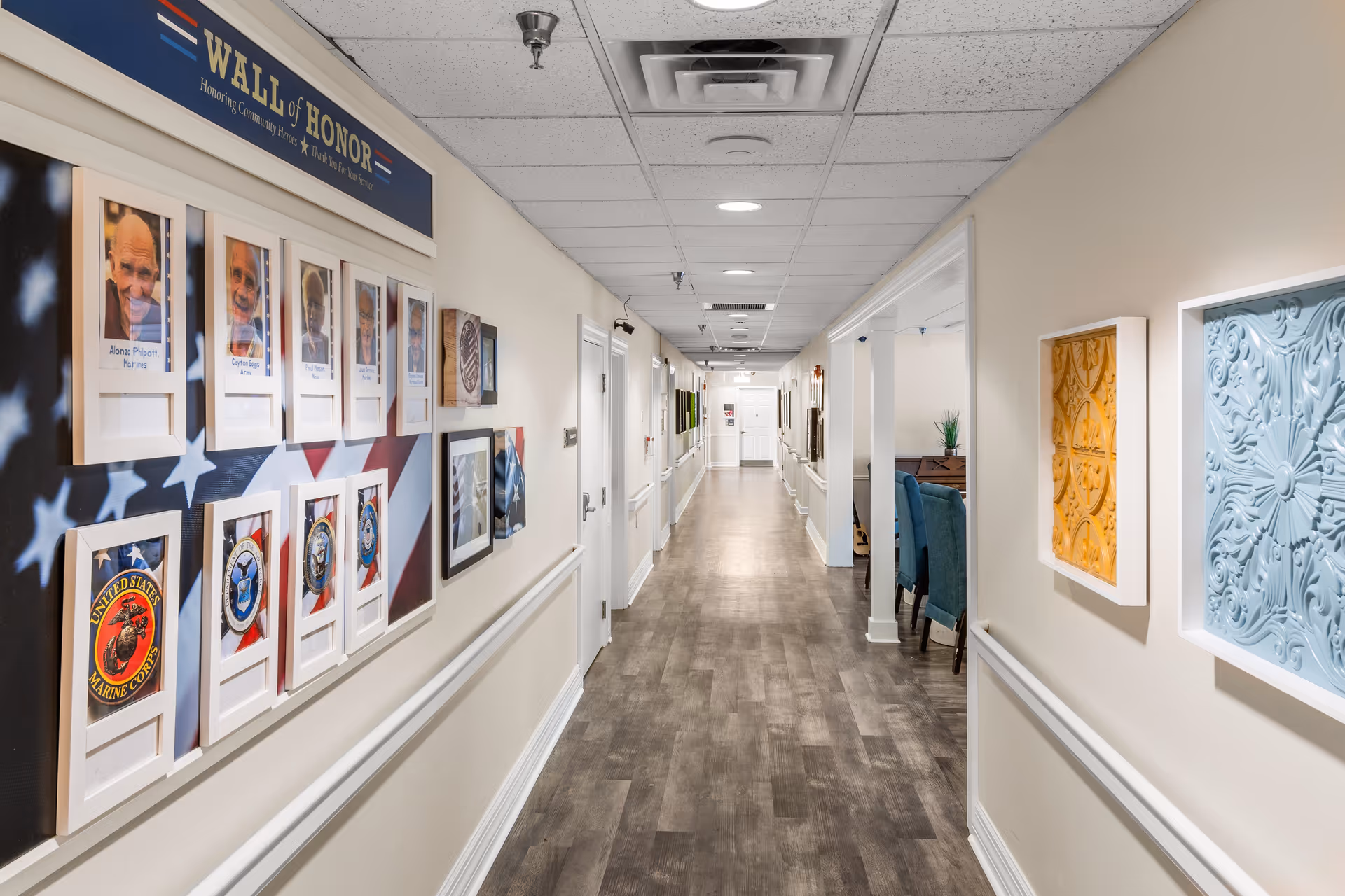 A long, well-lit hallway in a senior living facility with a 'Wall of Honor' display on the left wall featuring photos and military insignias. The right wall has decorative framed art pieces, and there is a seating area with blue chairs visible further down the hallway.