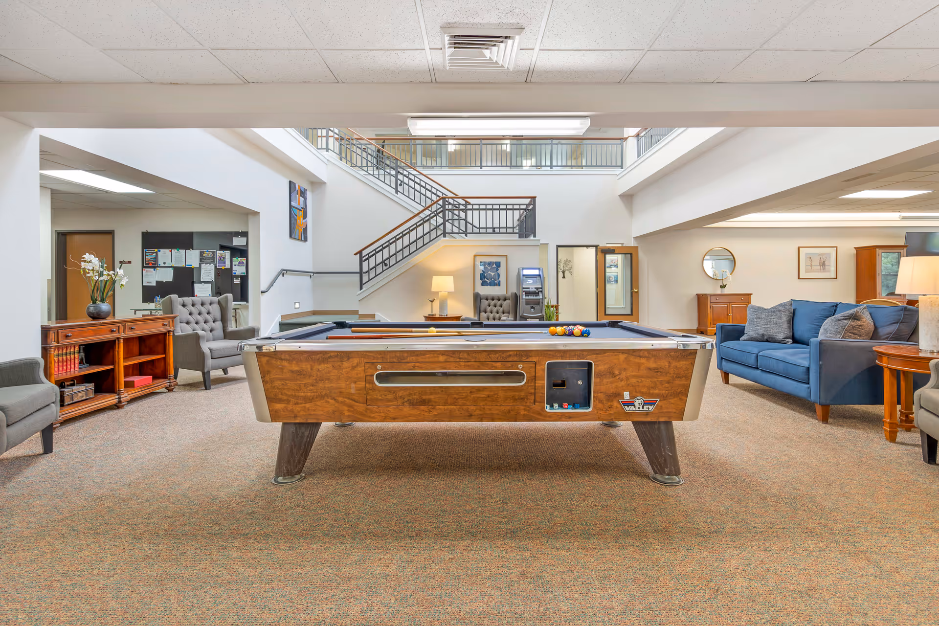 Interior view of a senior living facility common area featuring a pool table in the center, surrounded by comfortable seating including armchairs and a blue sofa. The space has a carpeted floor, a staircase with metal railings leading to an upper level, and various pieces of wooden furniture and decor such as a sideboard with a vase of flowers and framed artwork on the walls.