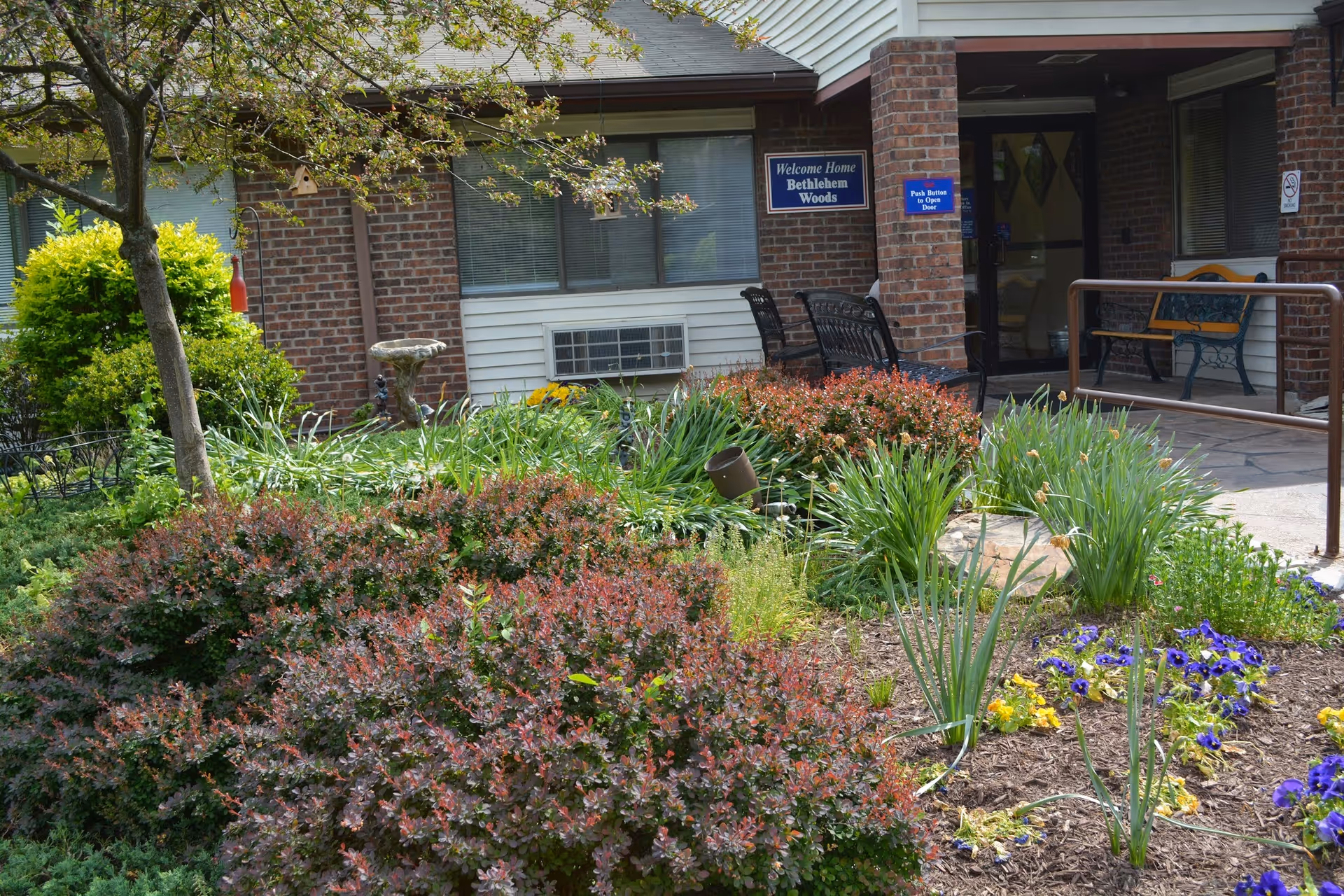 Entrance to Bethlehem Woods Nursing and Rehabilitation facility with a garden in the foreground featuring various plants and flowers. There are benches near the entrance and a sign that reads 'Welcome Home Bethlehem Woods'.