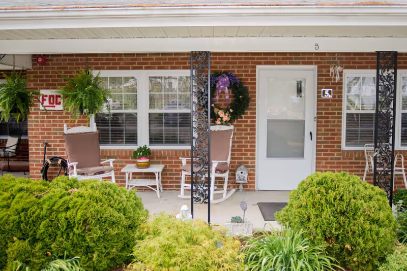 Front porch of a brick senior living unit with rocking chairs, potted plants, a wreath on the door, and shrubs in the foreground.