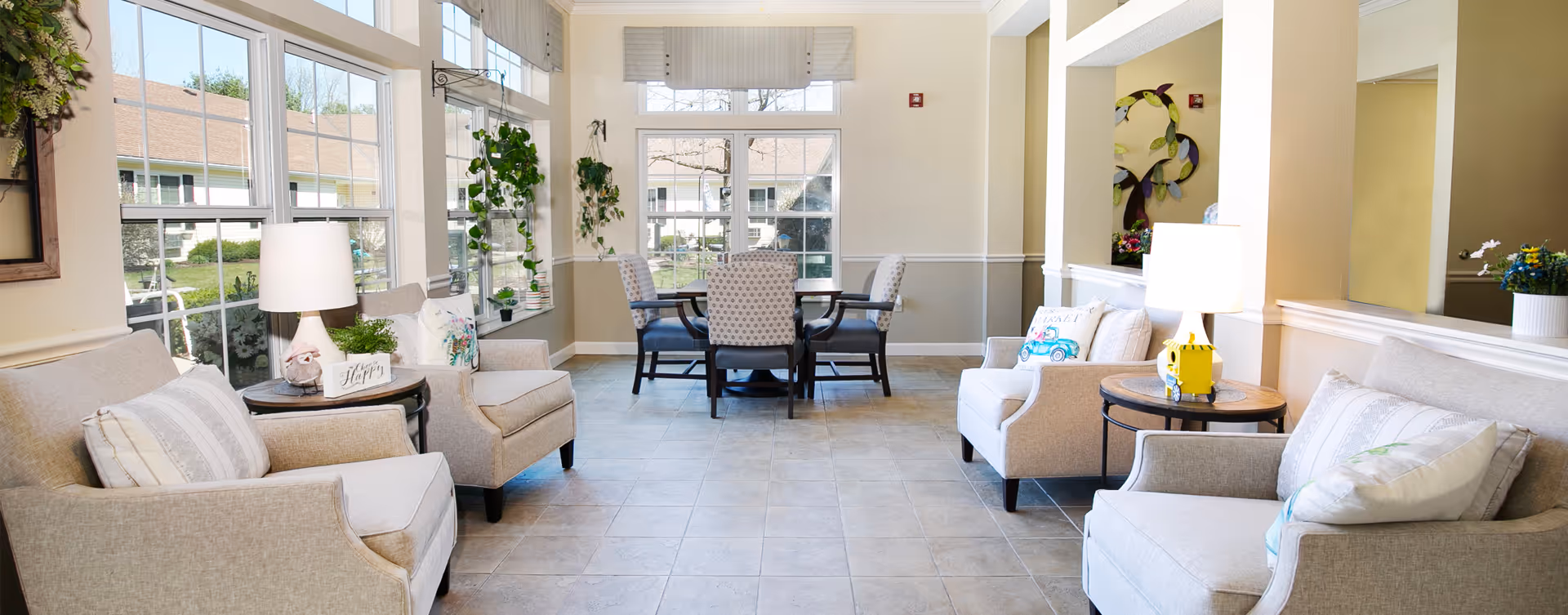 Bright communal sitting area with upholstered armchairs, side tables, and a dining table by large windows.