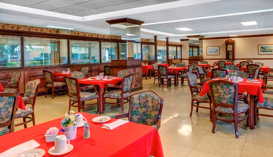 A spacious dining room with multiple tables covered in red tablecloths and floral-patterned chairs. Each table is set with white cups, plates, napkins, and small flower arrangements. The room features large windows along one wall, wooden paneling, and a grandfather clock in the background.