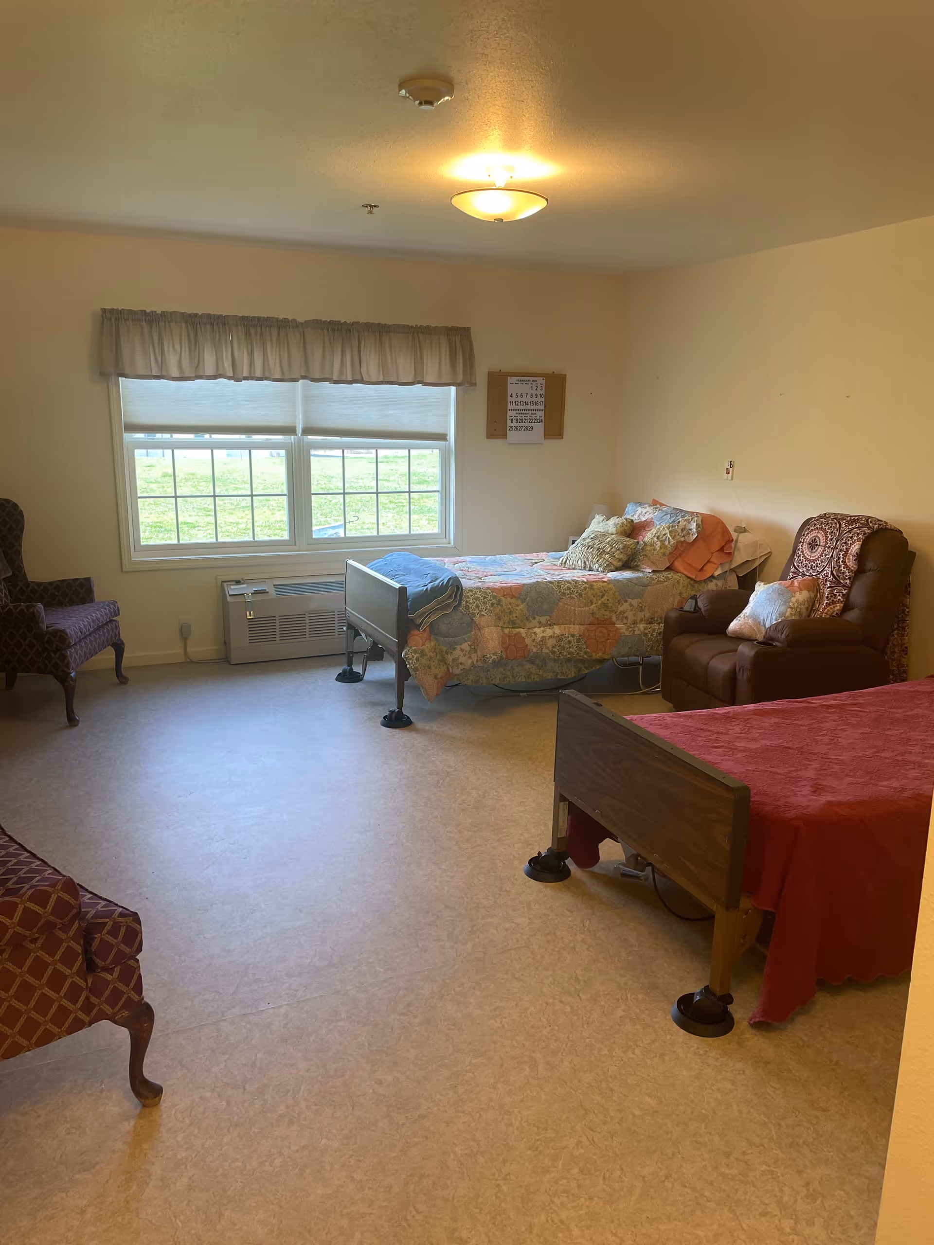 A bright assisted-living bedroom with two beds, recliner chairs, and a large window overlooking grass.