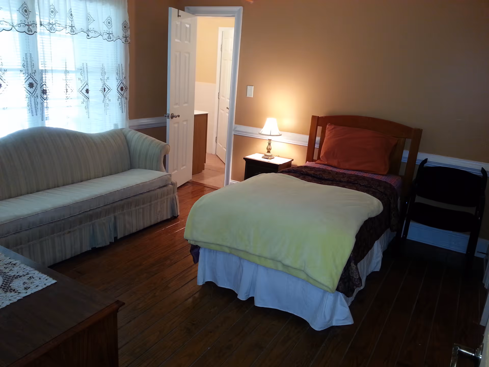 A cozy bedroom with a single bed covered with a yellow blanket and a brown pillow. Next to the bed is a small nightstand with a lit lamp. There is a striped sofa along the window with sheer curtains, and a wooden floor. An open door leads to a bathroom.