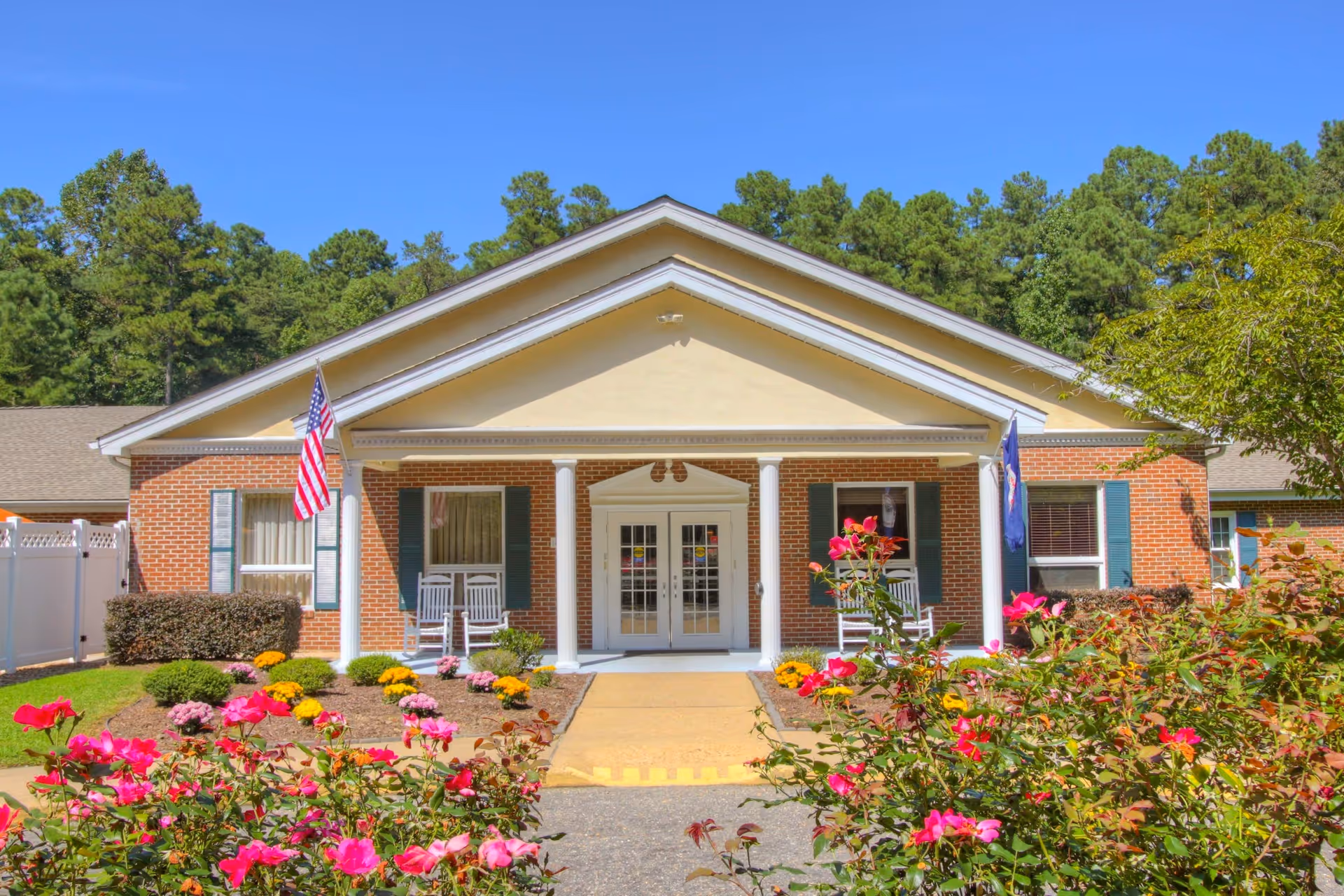 Brick single-story building entrance with columns, rocking chairs, flags, and colorful flower beds in front.