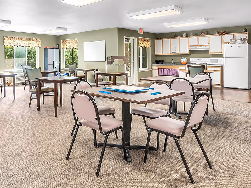 A bright room with several tables and chairs arranged for seating. The room has large windows with floral valances letting in natural light. In the background, there is a kitchenette area with white cabinets, a refrigerator, microwave, and a stove. The floor is carpeted and the walls are painted green.